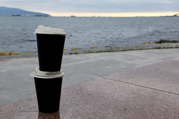 Two paper coffee cups stand on top of each other on a marble kerb against the blue sea. Takeaway cup with lid with ready drink. Holiday concept for two, recycling of used material