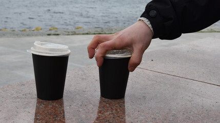 Two paper coffee cups stand on a marble border. A man's hand takes one glass of coffee against the background of the blue sea. Takeaway cup with lid with ready drink. Recycling of used material, recre