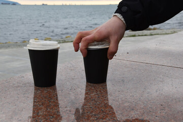 Two paper coffee cups stand on a marble border. A man's hand takes one glass of coffee against the background of the blue sea. Takeaway cup with lid with ready drink. Recycling of used material, recre