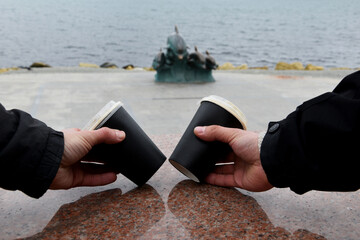 Two paper coffee cups stand on a marble border. Male hands take glasses of coffee against the background of the blue sea. Takeaway cup with lid with ready drink. Recycling of used material, recreation
