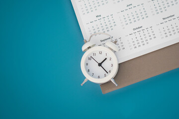 Fototapeta na wymiar white analog alarm clock on blurred yearly calendar with grunge blue background with copy space, business meeting schedule, travel planning or project milestone and reminder concept