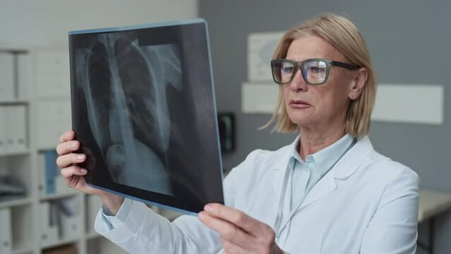 Serious mature female radiologist in eyeglasses and lab coat looking attentively at x-ray image of lungs of patient by her workplace while working in medical office