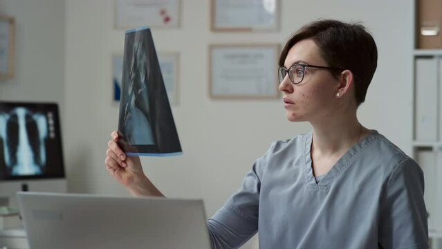 Young serious female radiologist or general practitioner in blue uniform comparing two x-ray images of her patients while sitting in front of laptop in clinics and gicing online consultations