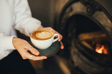 Close up female hands holding cup of hot cappuccino on wooden stool near burning fireplace.