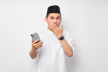 Surprised young Asian Muslim man holding mobile phone and covering mouth with hand isolated on white background. People religious Islam lifestyle concept. celebration Ramadan and ied Mubarak