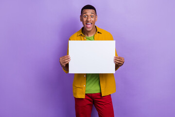 Photo of cheerful impressed guy dressed yellow shirt staring holding white banner announce sale isolated on violet color background