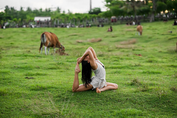 A beautiful young girl is doing yoga in nature, on the lawn, cows graze around.