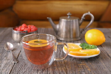 A glass cup of black tea with lemon and mint next to an old iron teapot and other tea accessories stands on a table against a log wall. 