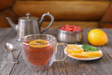 A glass cup of black tea with lemon and mint next to an old iron teapot and other tea accessories stands on a table against a log wall. 