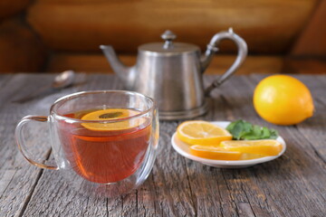 A glass cup of black tea with lemon and mint next to an old iron teapot and other tea accessories stands on a table against a log wall. 