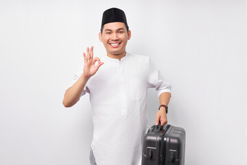Smiling young Asian Muslim man carrying a suitcase in hand and showing okay gesture isolated on white background. Ramadan and eid Mubarak concept