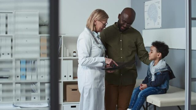 Young African American man and his cute little son listening to mature female radiologist in lab coat walking to them and explaining x-ray image with results of medical examination