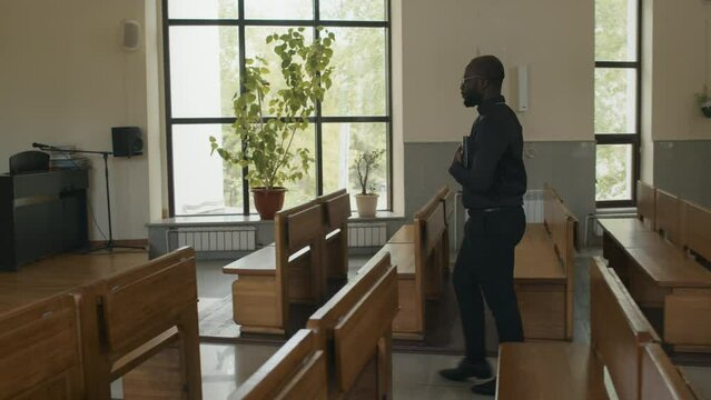 Long follow shot of young adult African American Catholic priest holding prayer book walking along church towards the altar