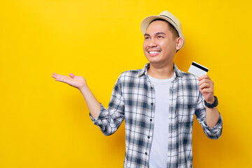 Cheerful Asian ethnicity young man 20s wearing casual clothes hat holding credit bank card, showing copy space with palm isolated on yellow background studio portrait. People lifestyle concept