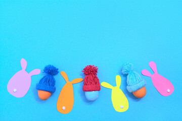 Painted Easter eggs in a winter knitted hat. Rabbit cut out of coloured paper. Blue background. Copy scape