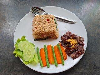 Fried rice, fried beef, vegetables, cucumbers, carrots, long beans, in a white plate on a wooden table.