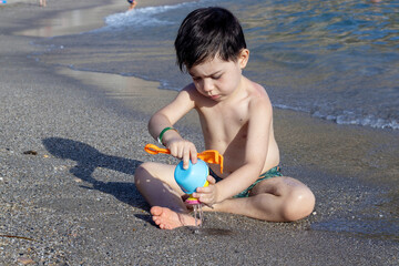 kid playing on beach sand running digging using plastic toys crab lobster seahorse shell shape rake and sprinkler.sunny summer vacation day child preschooler boy feet legs in water waves hitting sole