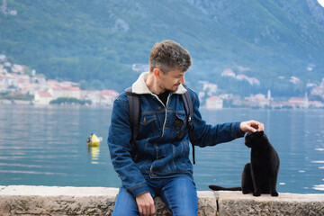 A cheerful guy (Caucasian race) sits on the embankment of the Bay of Kotor next to a black cat,...