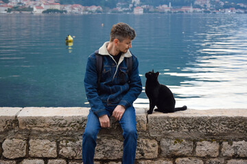 A cheerful guy (Caucasian race) sits on the embankment of the Bay of Kotor next to a black cat,...