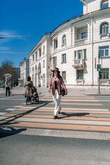 Woman city road crossing. Stylish woman in a hat crosses the road at a pedestrian crossing in the city. Dressed in white trousers and a jacket with a bag in her hands.