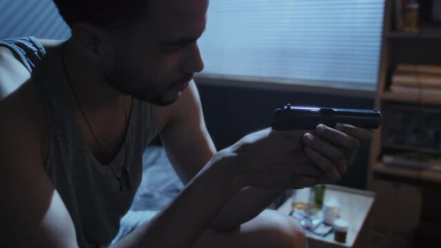 High angle handheld shot of young man with insomnia sitting on bed at night holding gun in hands thinking about traumatic events during military operation
