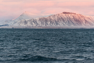 imagen de unas montañas iluminadas por las últimas luces del día, dando un tono magenta, con el cielo nublado y el mar en la parte inferior