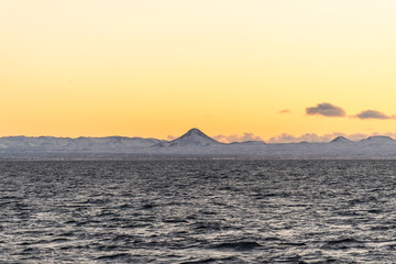 imagen del mar con una montaña de fondo y la puesta de sol iluminando el cielo con algunas nubes bajas 