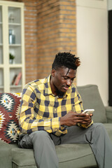 Handsome African man using smartphone while sitting on a sofa in cozy living room. Jpung man typing a message.