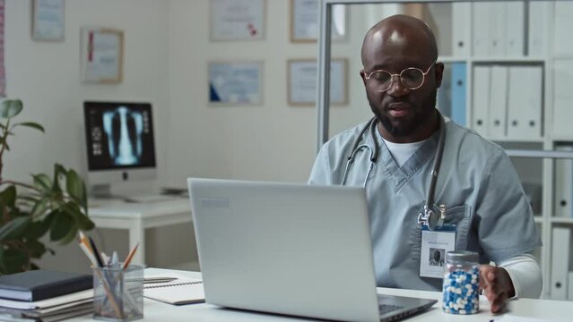 Young African American general practitioner in uniform and eyeglasses sitting by workplace in front of laptop during online consultation and recommending pills to patient
