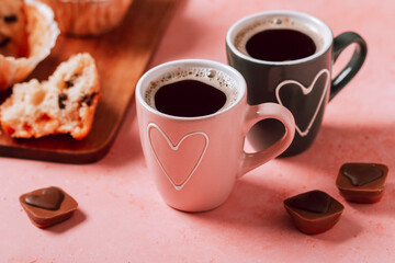 Two coffee cups with heart in pink and black colors, muffins and chocolates closeup