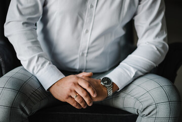 Fototapeta premium Male hands with wedding ring and watch. The man is sitting on the couch. Hands of the groom with an engagement ring. Close-up folded man's hands. Stylish man concept.