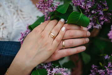 Fototapeta premium Rings. Engagement. Hands are newlyweds with wedding rings. Close up on the background of a wedding bouquet of flowers of lilac.