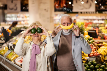 A silly senior couple is playing with fruits and making goofy faces at the supermarket. - Powered by Adobe