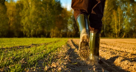 Tuinposter Close-up of a farmer's feet in rubber boots walking down a farmer field  dust rising from shoes. Low angle. One part is sown, the second part is not sown. © Valua Vitaly