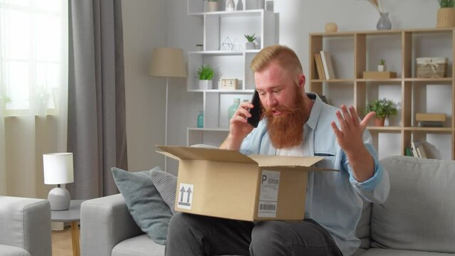 Redhead man sitting on sofa with opened parcel express complaints to seller or courier on the phone about damaged goods, crashed items inside cardboard box
