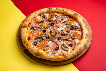 pizza with ham, tomatoes, mushrooms and olives on a board on a colored red-yellow background