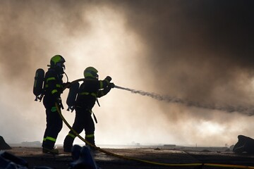 Silhouettes of firemen in smoke, using water to extinguish a massive fire in an industrial building
