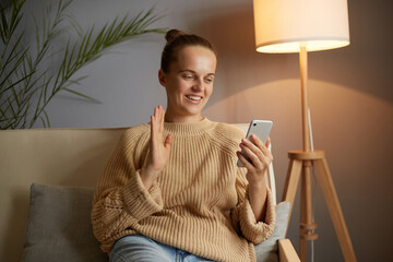 Indoor shot of adorable woman with bun hairstyle wearing beige sweater sitting on sofa in living...