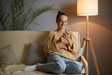 Photo of attractive smiling Caucasian woman wearing beige jumper and jeans sitting on cough, holding smart phone in hands, surfing internet, checking social networks.