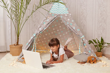 Indoor shot of dark haired little girl with pigtails lying in wigwam with laptop, watching cartoons or funny videos in internet, spending weekend in her cosy room. © sementsova321