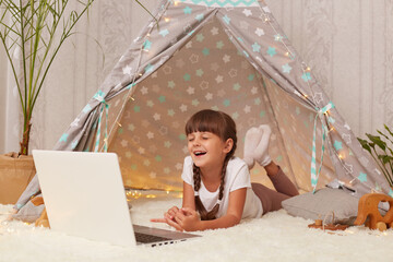 Portrait of laughing adorable cute little girl with pigtails lying in wigwam with laptop, spending time with computer, watching funny videos in internet. © sementsova321