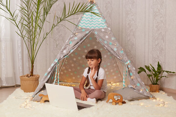 Portrait of joyful dark haired little girl with pigtails lying in peetee tent with laptop, having video call, waving hand, saying hello to her friend, looking at device screen. © sementsova321