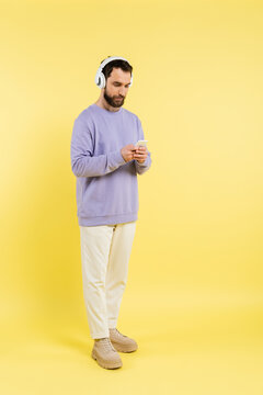 full length of trendy bearded man in wireless headphones using mobile phone on yellow.