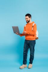 full length of happy student in orange hoodie and jeans using laptop on blue.