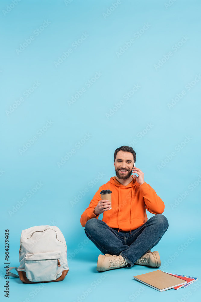 Sticker smiling student sitting with coffee to go and talking on smartphone near backpack and copybooks on blue.