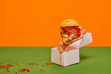 Food pop art photography. Female hand sticking out food box with burger on green tablecloth and orange background. Concept of taste, creativity, art. Complementary colors. Copy space for ad, text