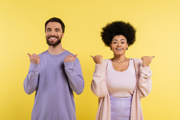 cheerful interracial couple pointing with thumbs and looking at camera isolated on yellow.