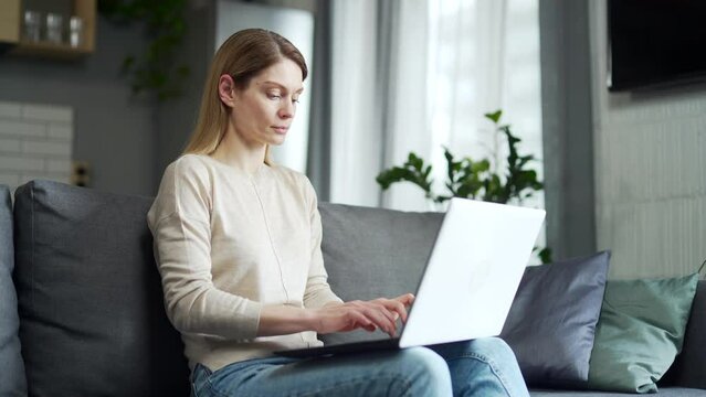 Serious caucasian woman using laptop while sitting on sofa in living room. Concentrated middle-aged female freelancer working at home, texting, checking email while holding device on lap1