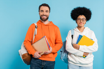 cheerful multiethnic students in hoodies with copybooks and backpacks looking at camera isolated on blue.