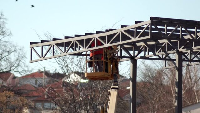 Workers standing on crane are welding metal beam outdoors at metal frame construction site